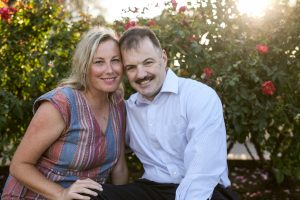 Image of couple sitting outside by foliage and smiling